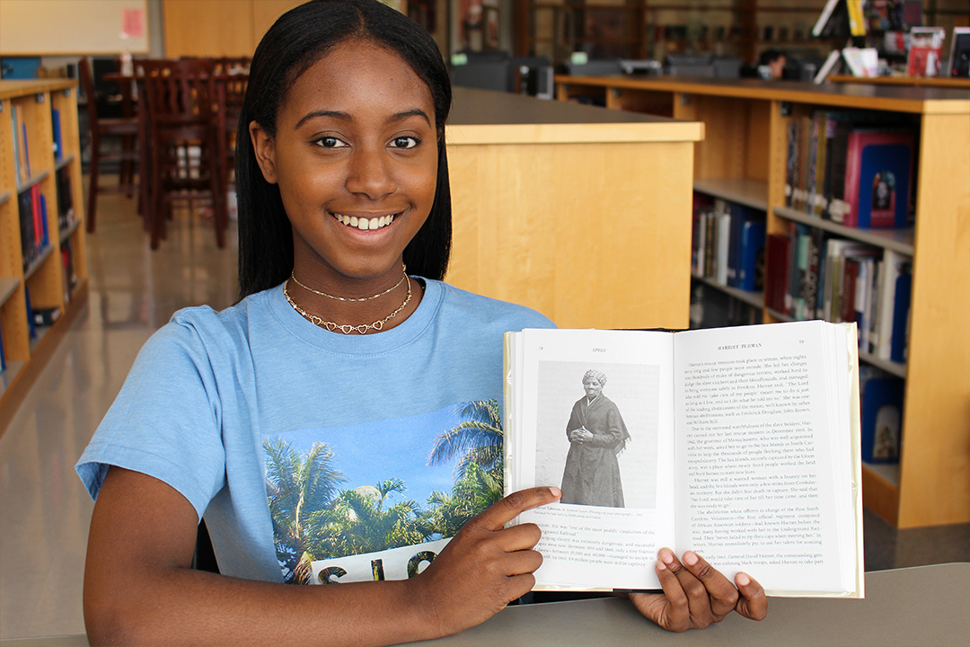 Jasmine sitting in a library pointing to a photo of Harriet Tubman in a book she is holding